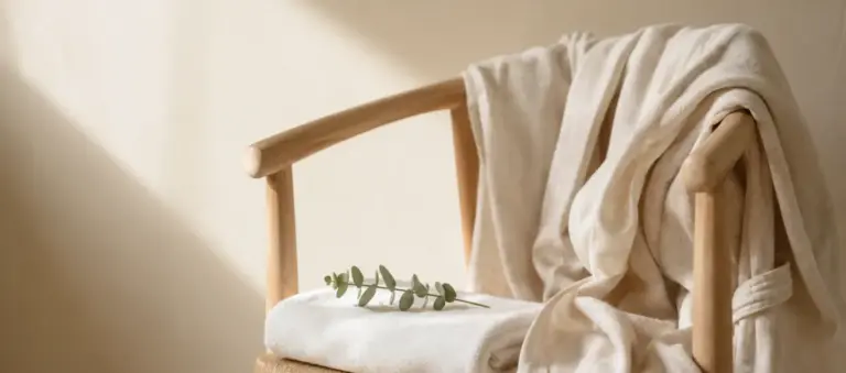 A cream linen robe on a wooden chair with a folded towel and a eucalyptus sprig